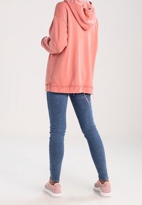 Růžová mikina s kapucí volného střihu, s třepeným lemem, spárovaná s modrými skinny džíny a světlými růžovými texturovanými botami.