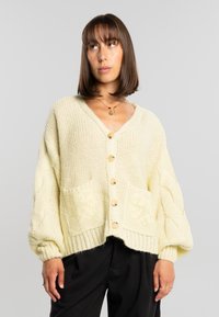 Mujer con cabello castaño hasta los hombros que lleva un cárdigan de punto crema de gran tamaño con bolsillos delanteros y botones de madera, combinado con pantalones negros.