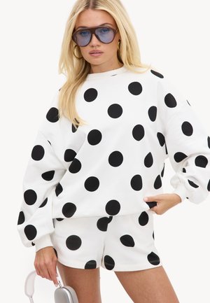 Femme blonde portant un sweat-shirt blanc oversize et un short à gros pois noirs, des lunettes de soleil teintées de bleu et des boucles d'oreilles en or en forme d'anneau.
