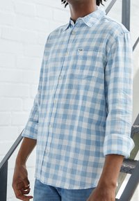 Ljusblå och vit rutiga skjorta med knappar, med en ficka, tillverkad av mjukt tyg, har långa ärmar som är uppkavlade, och en liten logotyp.