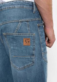 Jeans de denim azul con lavado claro, que presentan un gran bolsillo trasero y una etiqueta de cuero con un logo. Costuras distintivas a lo largo de las costuras.