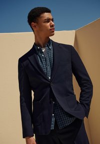 Marineblazer aus strukturiertem Stoff mit zwei frontalen Taschen, über einem blau karierten Hemd getragen. Das Modell steht vor einem hellbeigen Hintergrund.