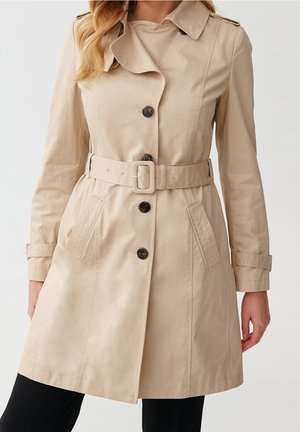 Trench beige avec des boutons noirs, une ceinture à la taille, des poches latérales et des pattes aux poignets, porté sur un pantalon noir.
