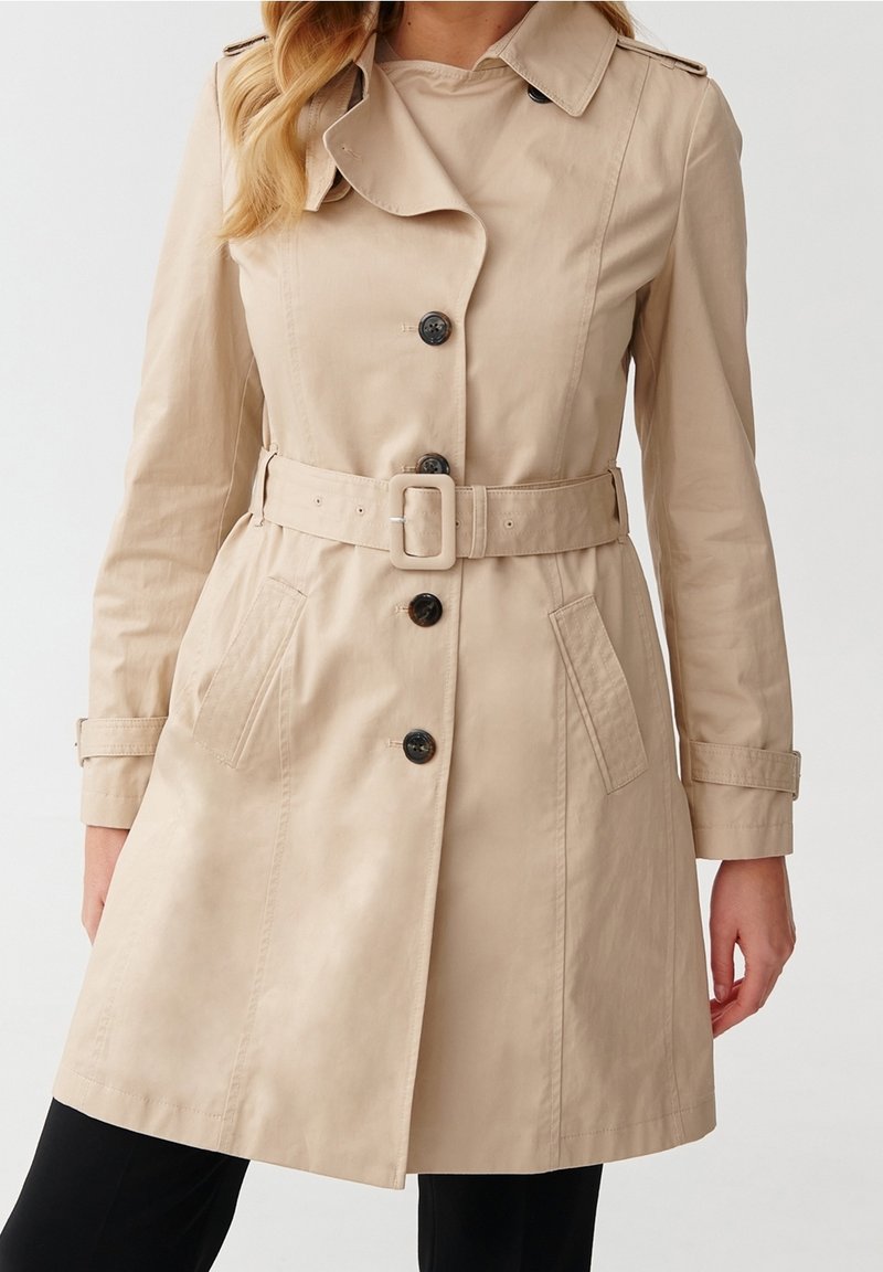 Trench beige avec des boutons noirs, une ceinture à la taille, des poches latérales et des pattes aux poignets, porté sur un pantalon noir.
