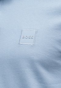 Υφασμα σε ανοιχτό μπλε χρώμα με υφή, που έχει ένα ραμμένο τετράγωνο πατς με την ανάγλυφη λέξη "BOSS" στο κέντρο.