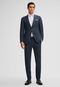 Donkerblauw op maat gemaakt pak van een gestructureerde stof, enkel knoopjasje, notch revers en slim-fit broek. Gestyleerd met een wit overhemd.