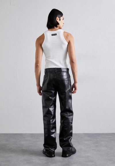 Μαύρα δερμάτινα παντελόνια με γυαλιστερή φινέτσα, συνδυασμένα με λευκό βαμβακερό αμάνικο μπλουζάκι. Ορατή ετικέτα μάρκας κοντά στον λαιμό. Απλό φόντο.