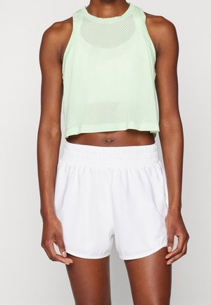 Personne athlétique portant un crop top en maille sans manches vert clair et un short blanc à taille élastique, debout devant un fond uni.