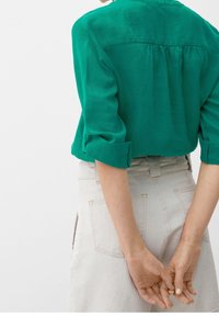 Grüne Bluse mit kurzen Ärmeln, Rückenpasse und Raffung, kombiniert mit hellen, hoch taillierten Hosen mit Gesäßtaschen.