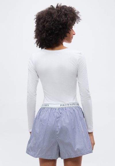 Personne aux cheveux bouclés portant une chemise blanche à manches longues et un short rayé bleu et blanc avec une ceinture Polo Ralph Lauren, vue de dos.