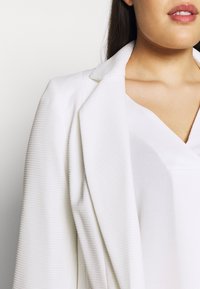 Bílý blazer s vertikálními žebrovanými vzory, límcovým límcem, zapínáním na jeden knoflík a splývavou siluetou, vrstvený přes bílou blůzu.