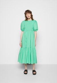 Vestido verde menta com corpete pregueado, mangas bufantes e saia em camadas. Usado com sandálias pretas de deslizar. Tecido suave e leve.