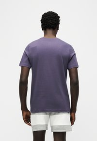 Άνδρας με κοντά σγουρά μαλλιά, φοράει απλό μοβ μπλουζάκι και σορτς με γκρι και λευκή διχρωμία, με την πλάτη στραμμένη σε λευκό φόντο.