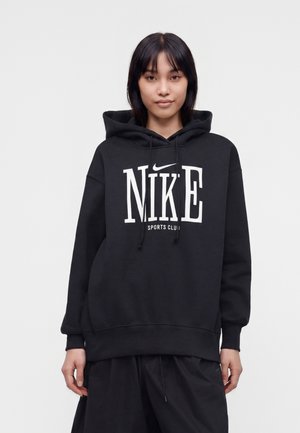 Ung kvinde med lige sort hår iført en sort Nike-hættetrøje med hvidt logo og tekst, stående mod en ensartet lys baggrund.