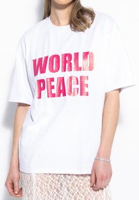 Biała bawełniana koszulka z krótkim rękawem, z wyrazistym tekstem "ŚWIATOWY POKÓJ" w różowym brokacie napisanym dużymi literami. Prosty, luźny krój.