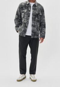 Mężczyzna w szarej, wzorzystej kurtce zapinanej na guziki, białej koszulce, czarnych spodniach i białych sneakersach z beżowymi i zielonymi podeszwami.