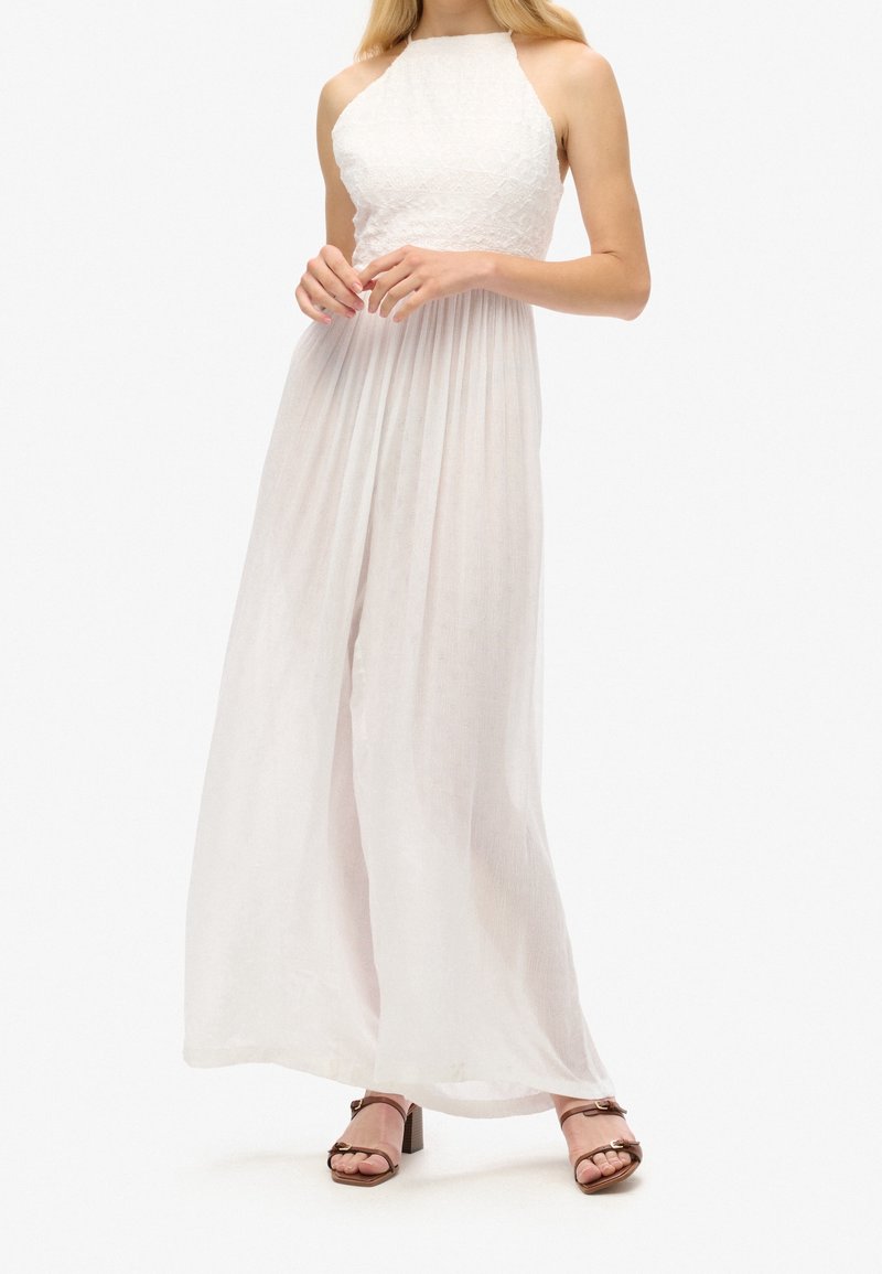 Femme portant une robe blanche en dentelle sans manches avec une jupe fluide et des sandales marron à talon bloc ouvert, debout devant un fond blanc.
