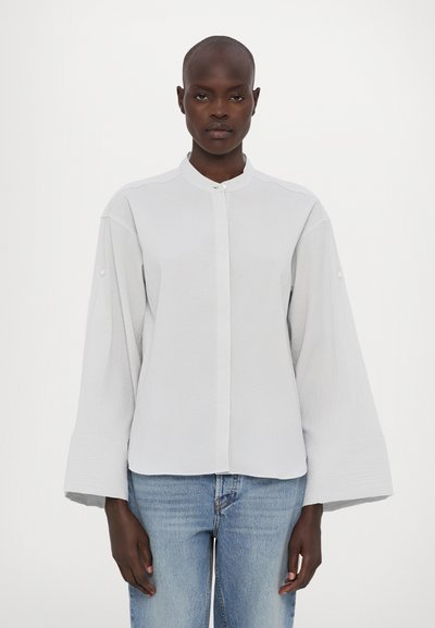 Chemise boutonnière gris clair avec col montant, tissu texturé, manches larges et détail de boutons subtils aux poignets, assortie à un jean bleu.