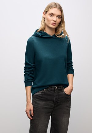 Felpa con cappuccio teal in morbido tessuto, caratterizzata da una vestibilità ampia e tasca a marsupio, abbinata a jeans grigi scuri.