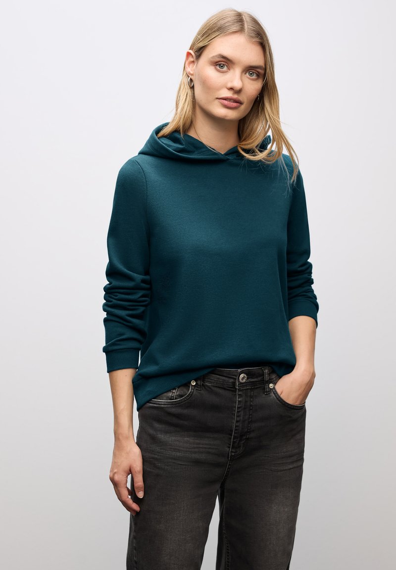 Felpa con cappuccio teal in morbido tessuto, caratterizzata da una vestibilità ampia e tasca a marsupio, abbinata a jeans grigi scuri.