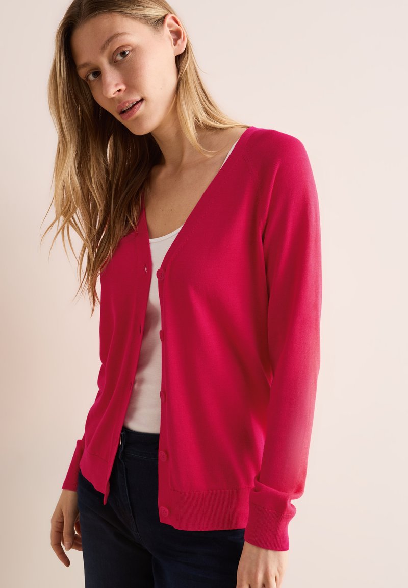 Cardigan fuchsia, à boutons sur le devant, manches longues, poignets et ourlet côtelés, matière légère, porté par-dessus un haut blanc.