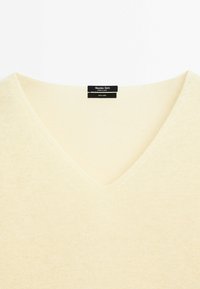Maglione in lino giallo chiaro con scollatura a V e lavorazione a texture. L'etichetta include il nome del marchio e i dettagli sul materiale.