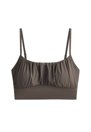 Femme debout portant un soutien-gorge de sport marron assorti et un legging taille haute avec des chaussures de sport noires sur un fond gris uni.
