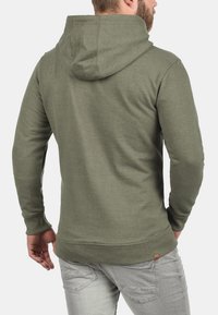 Olivgrüner Hoodie mit langen Ärmeln, einer Kordelzugkapuze und gerippten Bündchen. Enthält ein kleines Logo-Patch nahe dem Saum und hat eine weiche Textur.