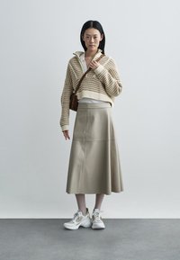 Nő áll fehér fal előtt, bézs kötött pulóvert, krémszínű midi szoknyát, fehér sportcipőt visel, és barna válltáskát hord.