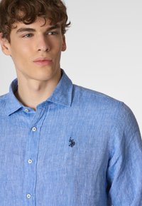 Camicia azzurra chiara con colletto in lino, caratterizzata da una superficie testurizzata. Presenta un logo ricamato nero sul lato sinistro del petto e bottoni bianchi.