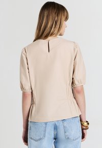 Blusa beige con maniche a sbuffo e dettaglio a chiave sulla schiena, lati arricciati e texture morbida. Abbinata a jeans in denim azzurro chiaro.