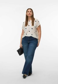 Vrouw met een wit T-shirt met zwarte hartjes, blauwe jeans met rechte pijpen, zwarte schoenen en een zwarte clutch in de hand, staand tegen een witte achtergrond.