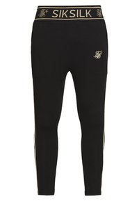 Černé legíny vyrobené z pružného materiálu s páskem zlaté barvy označeným "SIKSILK." Obsahují zlaté boční zipy pro dodatečný detail.