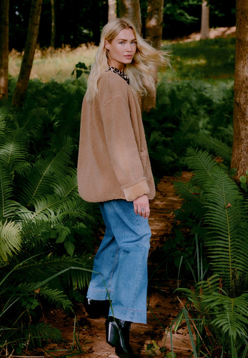 Beige fleecejakke med en løs pasform, parret med lyseblå brede jeans og sorte hæle støvler, sat blandt frodige grønne bregner.