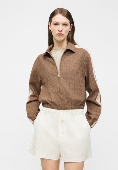 Γυναίκα με ίσια καστανά μαλλιά φοράει ένα καφέ μπουφάν με φερμουάρ και κρεμ ρίγες στα μανίκια, καθώς και αέρινα κρεμ σορτς, σε απλό φόντο.