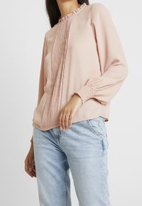 Blushrosa blus med volangkrage, vertikal plissering fram och samlade ärmslut. Tyget verkar slätt och lätt.