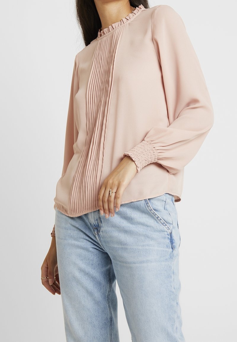 Blushrosa blus med volangkrage, vertikal plissering fram och samlade ärmslut. Tyget verkar slätt och lätt.