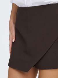 Primer plano de una persona que lleva una falda asimétrica marrón oscuro y una blusa blanca, con la mano apoyada en la cadera.