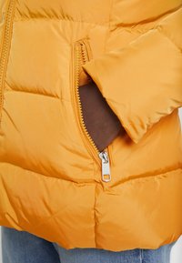 Hand i sidofickan på en ljusstark orange vadderad jacka, som synner del av blå jeans nedanför.