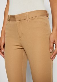 Pantalons ajustés beiges avec une texture lisse, une taille plate, des passants de ceinture et une poche frontale. Les coutures sont soigneusement piquées, offrant un look élégant.