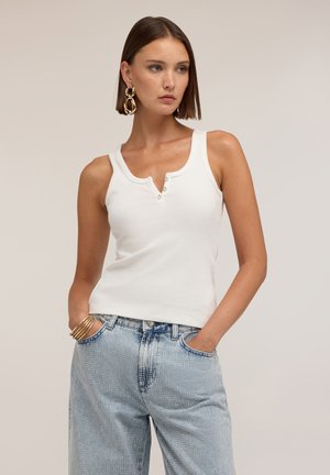 Mujer con el pelo corto y marrón, lleva una camiseta blanca sin mangas y vaqueros azules claros con tachuelas, manos en los bolsillos, mirando hacia un lado.