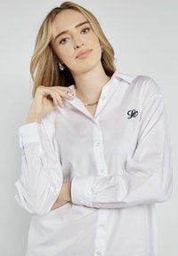 Biała koszula z długimi rękawami, wykonana z miękkiego materiału, z małym haftowanym logo na lewej piersi oraz klasycznym kołnierzykiem.