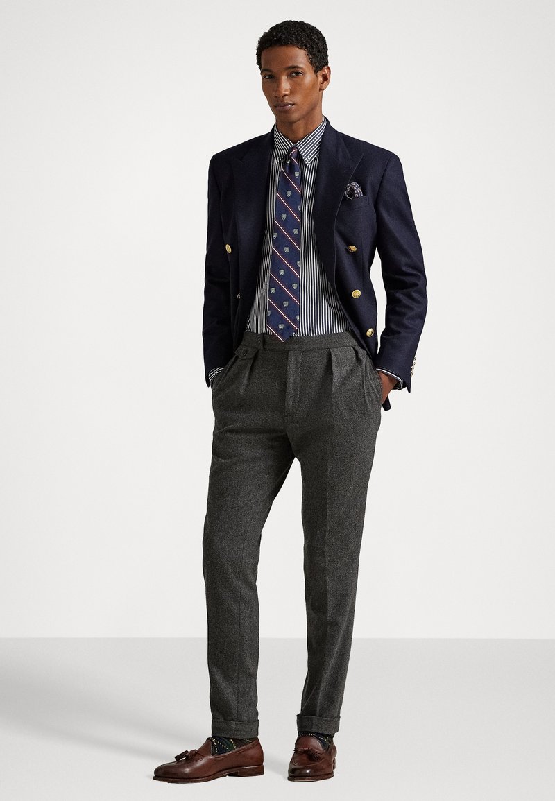 Blazer bleu marine avec boutons en or, chemise rayée, cravate à motifs, pantalon gris plissé et mocassins en cuir marron avec des chaussettes texturées.