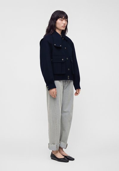 Femme debout portant une veste bleu marine foncé boutonnée, un jean gris clair retroussé et des ballerines noires sur un fond clair uni.