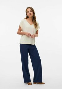 Haut v-neck crème à manches courtes, associé à un pantalon large bleu marine. La tenue présente un tissu lisse et un design simple. Chaussures plates marron.