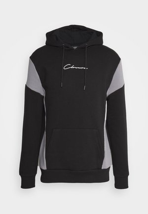 Hoodie noir avec des panneaux latéraux gris, des cordons de serrage et une poche réchauffe-mains à l'avant. Présente un logo signature blanc sur la poitrine. Texture douce.