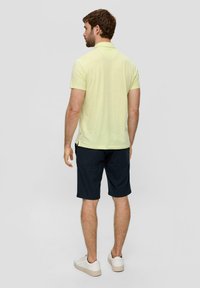 Svijetložuta polo majica s kratkim rukavima, u kombinaciji s tamnoplavim šortsevima. Model nosi bijele tenisice. Pogled s leđa, s jednostavnom pozadinom.