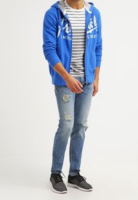 Hombre vestido con una sudadera azul con cremallera, camiseta de rayas grises y blancas, jeans claros rasgados y zapatillas negras de pie contra un fondo blanco.
