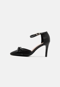 Black leather high-heeled shoe with pointed toe, ankle strap and gold buckle, featuring a glossy bow detail on the vamp.