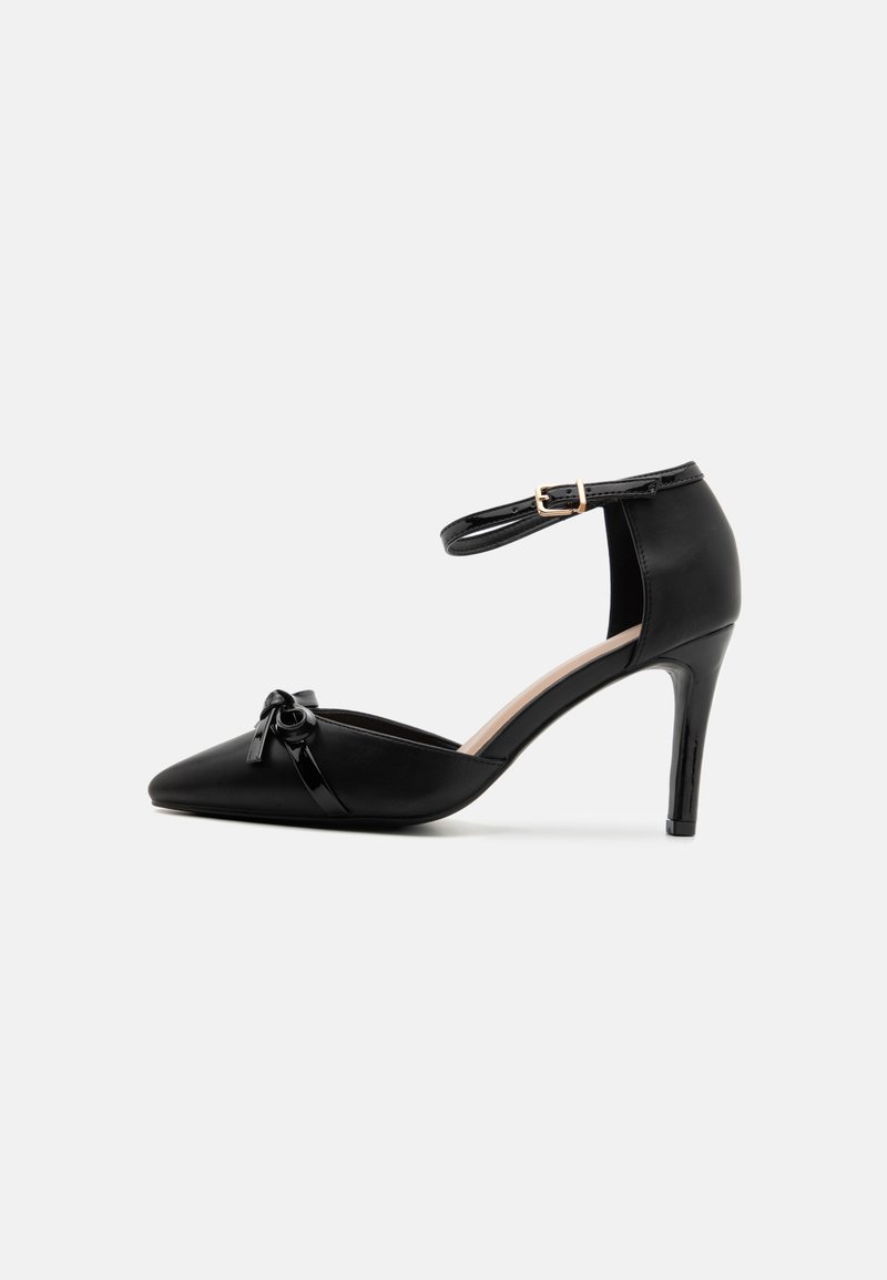 Black leather high-heeled shoe with pointed toe, ankle strap and gold buckle, featuring a glossy bow detail on the vamp.